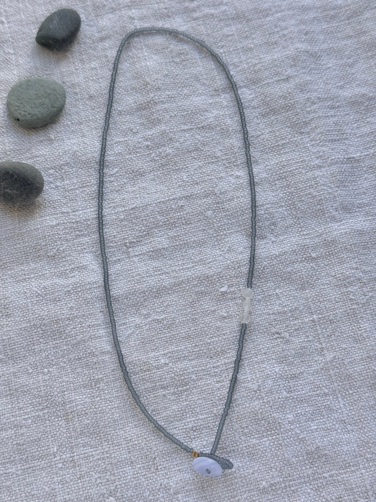 Collier lasso Jour de Pluie en entier, perles gris translucide mat, pierres de lune et calcédoine bleue, posé sur du lin avec des galets — fait main en Normandie
