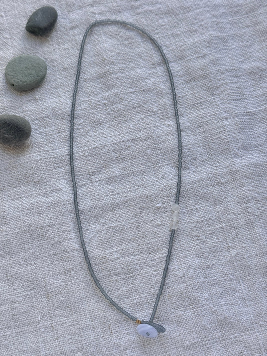 Collier lasso Jour de Pluie en entier, perles gris translucide mat, pierres de lune et calcédoine bleue, posé sur du lin avec des galets — fait main en Normandie