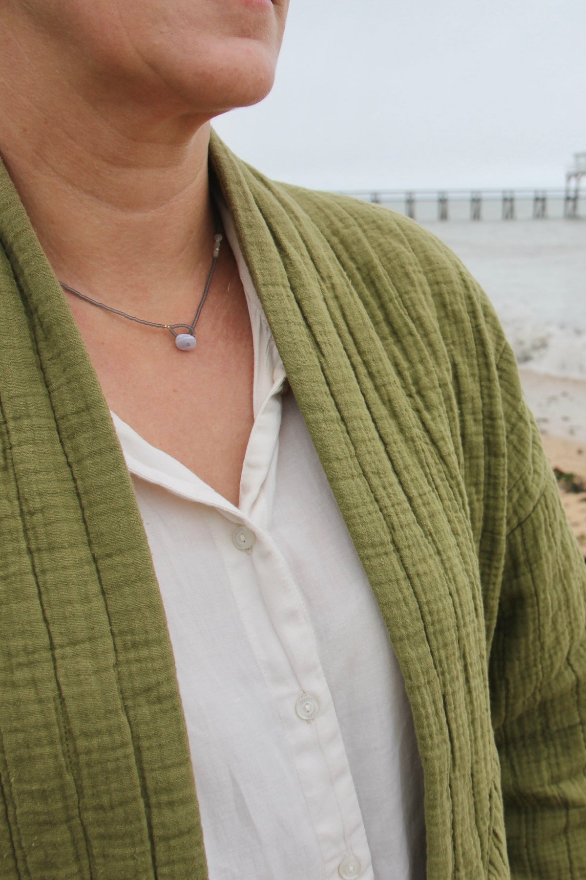 Collier lasso Jour de Pluie porté face à la mer, avec une veste vert olive et une chemise blanche