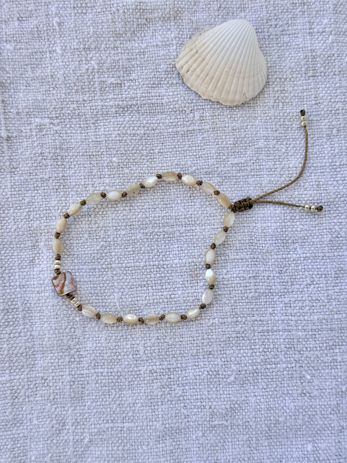 Bracelet Lichen en entier, nacres beige nouées sur cordon olive et carré de nacre abalone, posé sur du lin avec un coquillage — fait main en Normandie