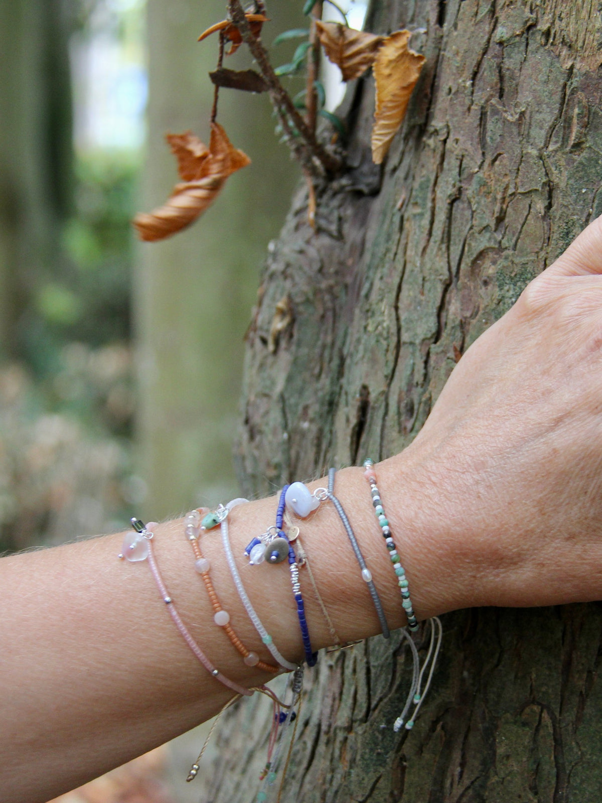 Accumulation de bracelets By Petite Pomme portés au poignet contre un arbre, perles et pierres fines en rose, bleu, gris et turquoise